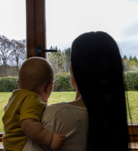 Photo taken from behind of mum with long dark hair holding baby wearing mustard yellow top while looking out of window.
